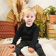 Raphaël a rejoint le concours — aidez-le/la à gagner de superbes lots ! toddler, child, blonde_hair, black_clothing, barefoot, pillows, floor, plants, pottery, wooden_crate, home_decor, pampas_grass, indoor, sitting, portrait, cute, cozy, boho_style, flooring, expression
