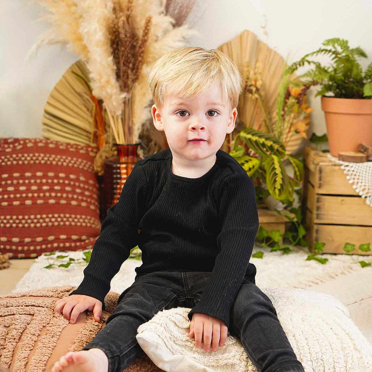 Raphaël a rejoint le concours — aidez-le/la à gagner de superbes lots ! barefoot, black_clothing, blonde_hair, boho_style, child, cozy, cute, expression, floor, flooring, home_decor, indoor, pampas_grass, pillows, plants, portrait, pottery, sitting, toddler, wooden_crate