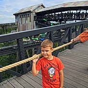 Valentin participe au concours pour gagner de l'argent avec cette photo : child, boy, orange_tshirt, rope, bridge, wooden_floor, building, people, crowd, outdoor, daytime, sky, casual_clothing, curious, holding, portrait, person, jeans, short_hair, fence