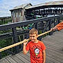 child, boy, orange_tshirt, rope, bridge, wooden_floor, building, people, crowd, outdoor, daytime, sky, casual_clothing, curious, holding, portrait, person, jeans, short_hair, fence