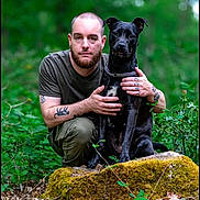 Ava participe au concours pour gagner de l'argent avec cette photo : man, dog, black_dog, tattoos, forest, greenery, moss, rock, outdoor, nature, pet, animal, portrait, crouching, casual_clothing, loyalty, companionship, beard, short_hair, calm