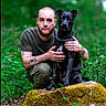 animal, beard, black_dog, calm, casual_clothing, companionship, crouching, dog, forest, greenery, loyalty, man, moss, nature, outdoor, pet, portrait, rock, short_hair, tattoos