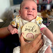 Esmae is registered to the contest to win money with this photo: baby, bee_pattern, blanket, blurred_background, child, cute, face, furniture, hand, happy, holding, indoor, infant, onesie, person, portrait, rainbow, ring, smile, wooden_sign