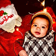 Khylae joined the competition — help win amazing prizes! baby, child, face, hat, argyle_sweater, red_clothing, blanket, christmas, holiday_lights, festive, smiling, indoors, cute, infant, portrait, cozy, toy, decoration, warm_lighting, holiday_theme