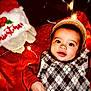baby, child, face, hat, argyle_sweater, red_clothing, blanket, christmas, holiday_lights, festive, smiling, indoors, cute, infant, portrait, cozy, toy, decoration, warm_lighting, holiday_theme