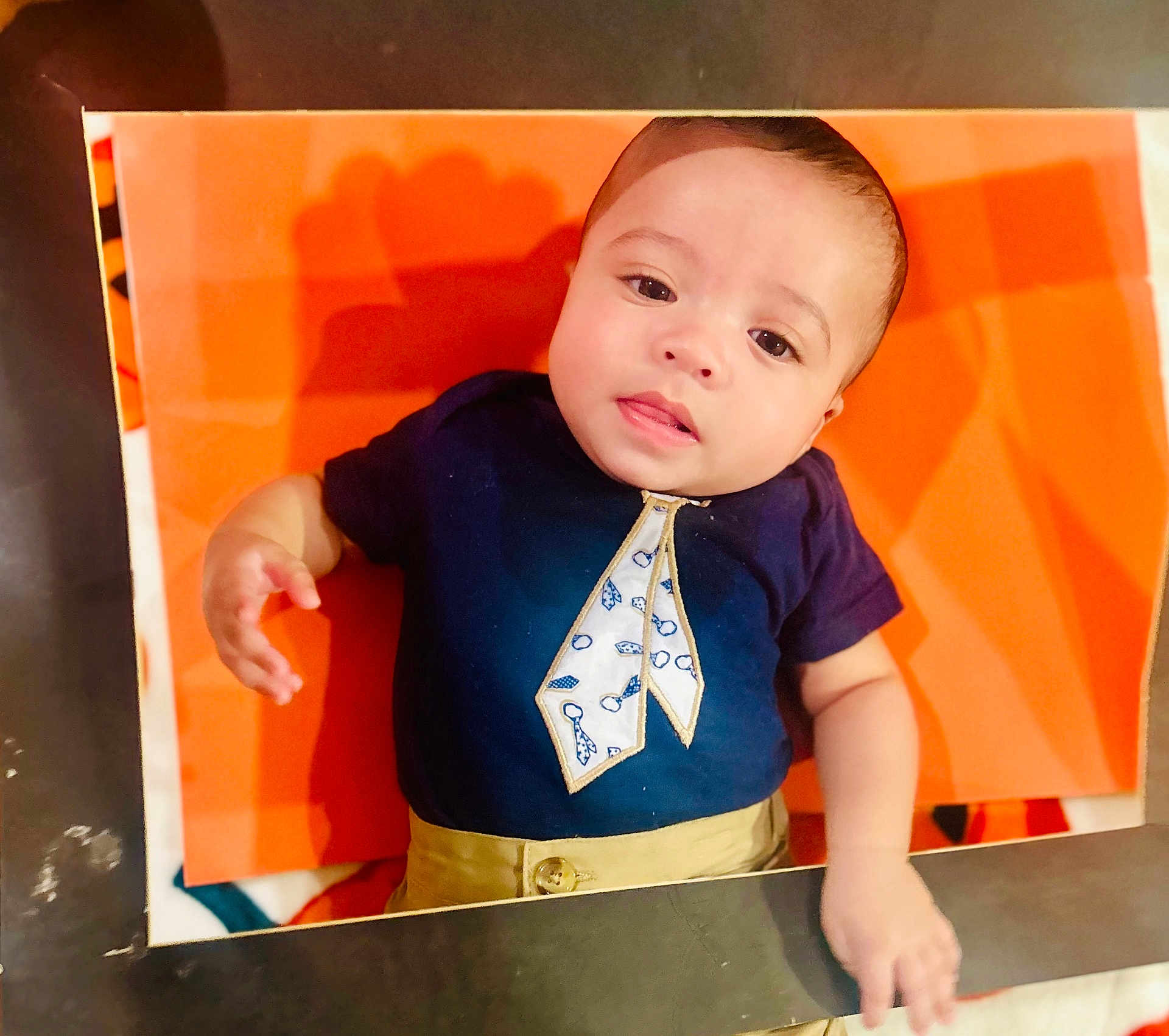 Khylae joined the competition — help win amazing prizes! baby, infant, child, orange_background, navy_blue_shirt, tie_design, khaki_pants, cute, portrait, face, eyes, hand, lying_down, clothing, expression, indoor, photo_frame, skin, curious, young