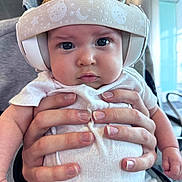 Ariella joined the competition — help win amazing prizes! baby, headphones, hands, person, clothing, face, expression, indoor, infant, cute, chubby_cheeks, white_onesie, holding, closeup, headband, pattern, blurred_background, human, portrait, serious_expression