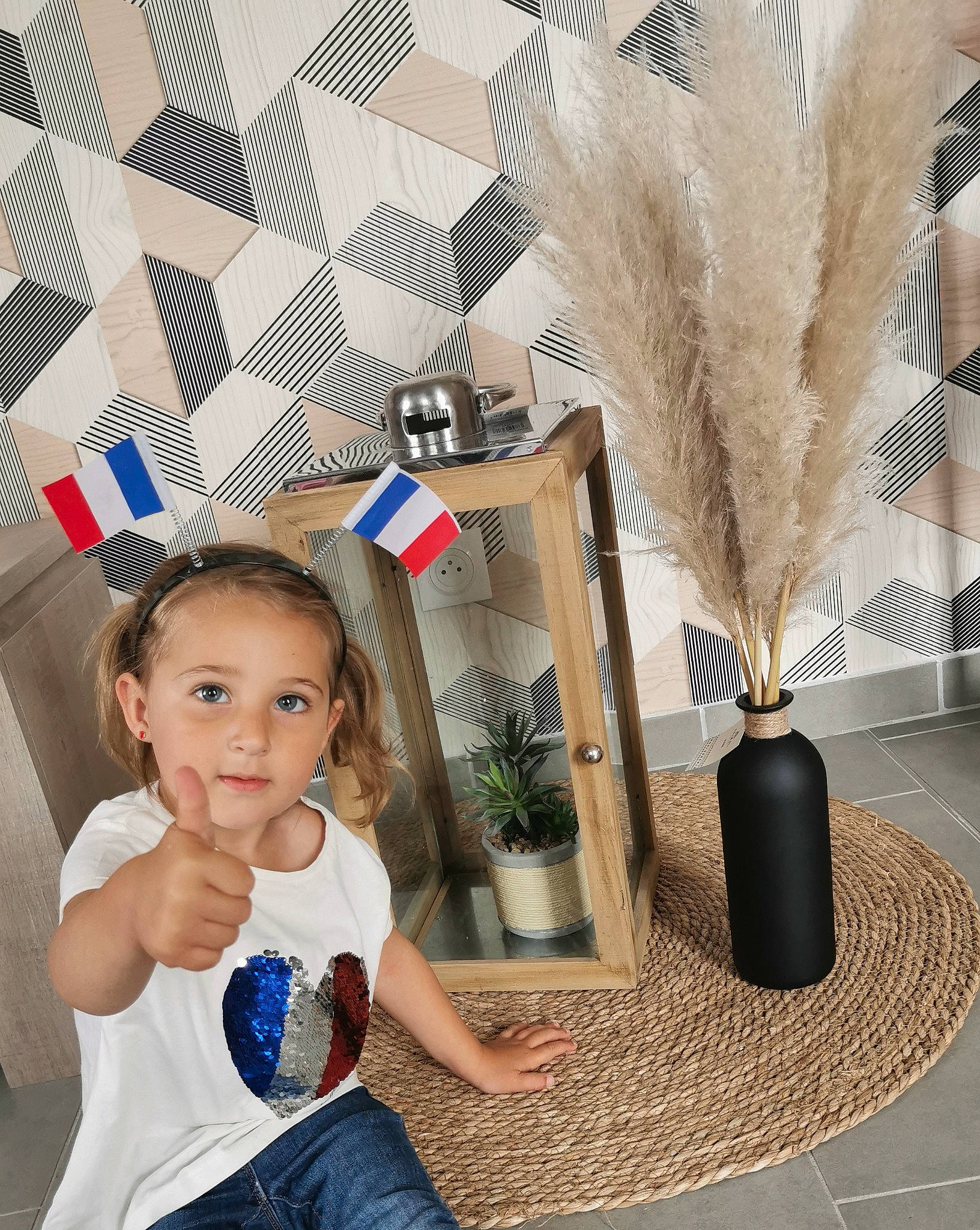 Zélie participe au concours pour gagner de l'argent avec cette photo : baby_toddler_clothing, blue, feather, floor, flooring, flowerpot, hairstyle, happy, houseplant, interior_design, person, photograph, plant, standing, t_shirt, toddler, twig, vertebrate, wall, white