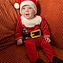 baby, child, santa_hat, christmas_outfit, red_clothing, couch, orange_couch, smiling, happy, indoors, holiday, festive, cute, infant, person, seated, plush, cozy, portrait, young_child