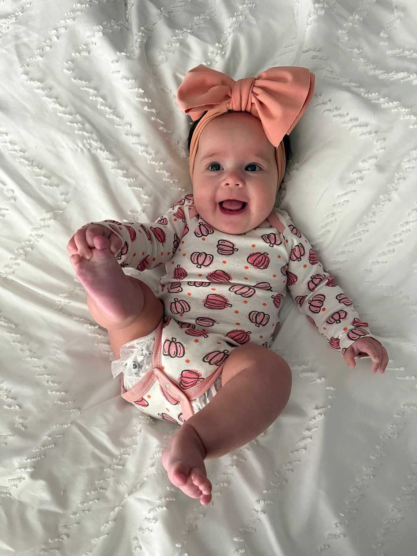 Nyra is registered to the contest to win money with this photo: baby, infant, smiling, headband, pink_bow, onesie, pumpkin_pattern, barefoot, lying_down, white_blanket, happy, cute, child, skin, foot, hand, face, playful, indoors, portrait
