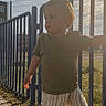 Raphaël a rejoint le concours — aidez-le/la à gagner de superbes lots ! child, toddler, outdoor, sunlight, fence, blue_fence, striped_pants, green_shirt, white_shoes, grass, sidewalk, shadow, daylight, sky, clouds, person, standing, casual_clothing, portrait, urban