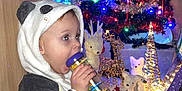 Raphaël a rejoint le concours — aidez-le/la à gagner de superbes lots ! toddler, child, panda_onesie, toy, colorful, christmas_tree, lights, ornaments, santa_figure, reindeer_decorations, presents, floor, holiday, festive, indoors, cozy, cute, sitting, playful, warm_lighting