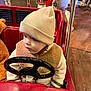 Raphaël a rejoint le concours — aidez-le/la à gagner de superbes lots ! toddler, child, hat, jacket, toy_car, steering_wheel, indoor, carousel, amusement_ride, floor, wooden_floor, red, beige, curious, cozy, clothing, face, person, play, entertainment
