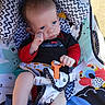baby, infant, child, stroller, seated, portrait, face, blue_eyes, wide_eyed, looking_at_camera, red_sleeves, black_clothing, harness, blanket, patterned_fabric, cartoon_animals, outdoor, sunlight, baby_gear, colorful