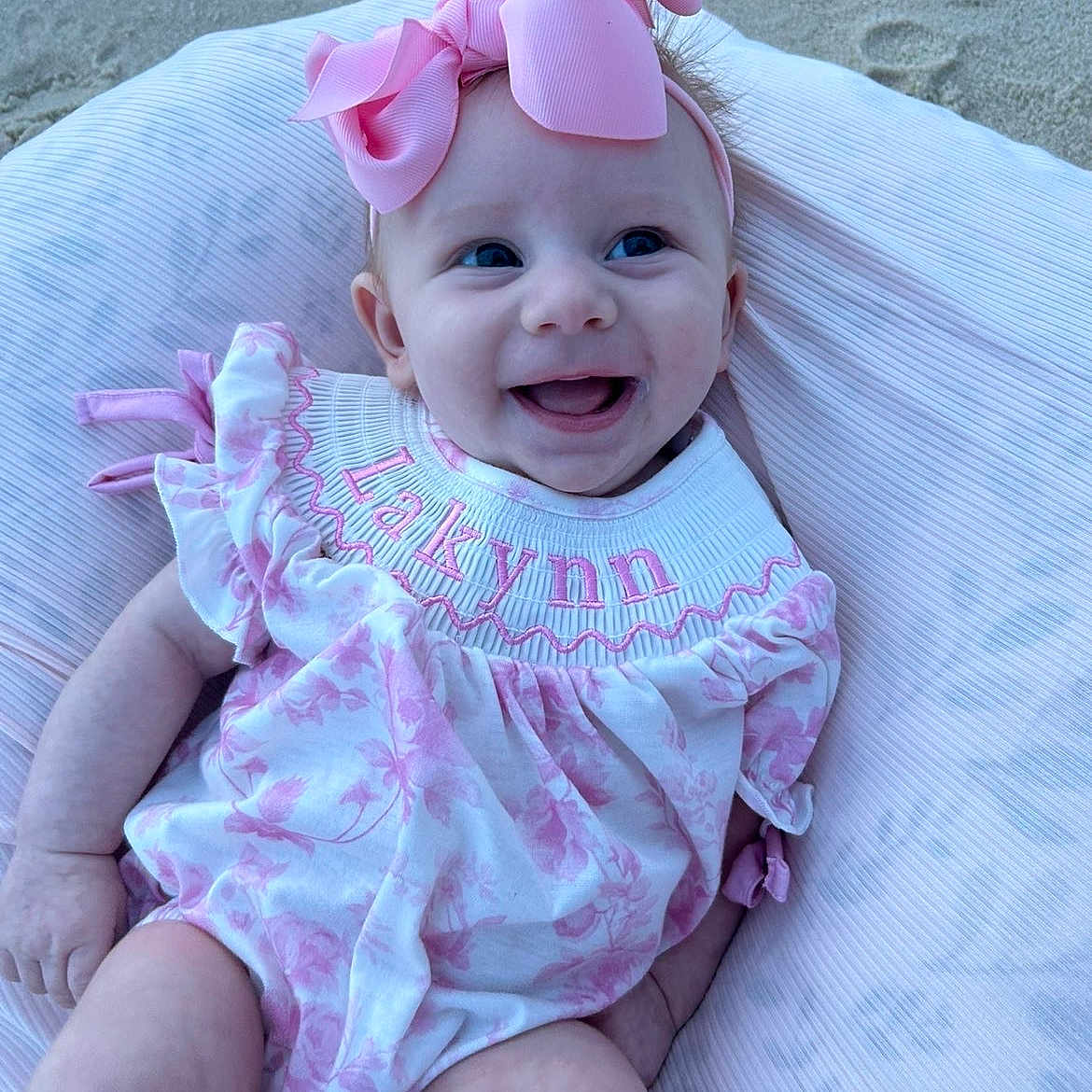 Lakynn is registered to the contest to win money with this photo: baby, infant, smile, pink_bow, floral_dress, pillow, sand, happy, cute, child, person, outdoor, portrait, headband, clothing, baby_girl, eyes, skin, face, babywear