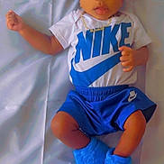 Messiah is registered to the contest to win money with this photo: baby, infant, child, blue_beanie, blue_booties, nike_shorts, nike_tshirt, gold_necklace, lying_down, curious_expression, soft_surface, casual_clothing, cute, portrait, indoor, young_child, fashion, footwear, headwear, person