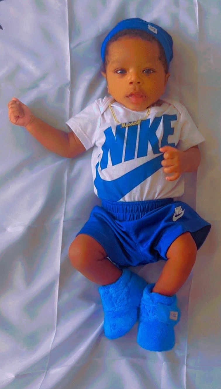 Messiah is registered to the contest to win money with this photo: baby, infant, child, blue_beanie, blue_booties, nike_shorts, nike_tshirt, gold_necklace, lying_down, curious_expression, soft_surface, casual_clothing, cute, portrait, indoor, young_child, fashion, footwear, headwear, person