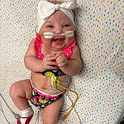 Serenity is registered to the contest to win money with this photo: baby, blanket, bow, cheerful, child, colorful, cute, face, hands, happy, headband, hospital, infant, lying_down, medical_tubes, patterned, skin, smiling, socks, swimsuit