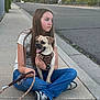 girl, dog, sidewalk, street, leash, harness, casual_clothing, long_hair, sneakers, trees, residential_area, sunset, outdoor, pet, animal, human, seated, calm, suburb, pavement