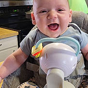 Mccoy is registered to the contest to win money with this photo: baby, infant, smiling, high_chair, breast_pump, kitchen, adult, man, cabinet, appliance, countertop, clothing, face, person, happy, indoor, furniture, table, sitting, parent