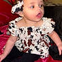 Iris is registered to the contest to win money with this photo: baby, child, headband, dress, cow_print, sitting, red_bedspread, black_pants, grey_socks, indoor, person, cute, young_child, portrait, fashion, expression, curious, skin, face, hair