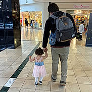 Iris is registered to the contest to win money with this photo: mall, shopping_mall, adult, toddler, child, walking, holding_hands, backpack, floor_tiles, storefront, people, indoor, casual_clothing, curly_hair, dress, white_tights, sneakers, store_sign, shopping, companionship