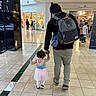 mall, shopping_mall, adult, toddler, child, walking, holding_hands, backpack, floor_tiles, storefront, people, indoor, casual_clothing, curly_hair, dress, white_tights, sneakers, store_sign, shopping, companionship