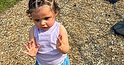 Iris is registered to the contest to win money with this photo: toddler, child, outdoor, playground, grass, wood_chips, braided_hair, flower, purple_top, blue_shorts, pink_shoes, sunny_day, motion, young_child, casual_clothing, curious_expression, small_hands, grass_field, play_area, summer_clothing