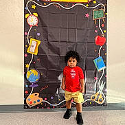 Josiah joined the competition — help win amazing prizes! toddler, child, boy, standing, red_shirt, yellow_shorts, black_shoes, curly_hair, school_backdrop, illustrations, clock, globe, apple, scissors, paint_palette, chalkboard, floor, wall, indoor, portrait