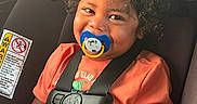 Josiah is registered to the contest to win money with this photo: toddler, child, car_seat, pacifier, curly_hair, orange_shirt, seatbelt, smiling, happy, indoors, safety, baby, person, portrait, young_child, car_interior, child_safety, seatbelt_buckle, cute, secure