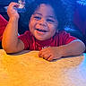 child, toddler, curly_hair, smiling, happy, red_shirt, table, indoor, portrait, face, person, cute, sitting, furniture, bright_lighting, skin, arm, hand, background, cozy