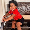 child, toddler, smiling, curly_hair, portrait, seated, bench, wooden_bench, red_shirt, black_shorts, sneakers, outdoor, wall, ornate_design, happy, looking_away, curious, casual_clothing, playful, photograph