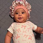 Legacy is registered to the contest to win money with this photo: baby, infant, smiling, pink_bonnet, onesie, bows, quilted_blanket, lying_down, cute, face, child, adorable, happy, indoors, portrait, person, small_child, headwear, fabric, soft_lighting