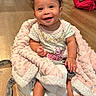 baby, child, smiling, blanket, pink, curly_hair, baby_seat, floor, wooden_floor, indoor, cute, happy, person, infant, clothing, foot, hand, backpack, toy, portrait
