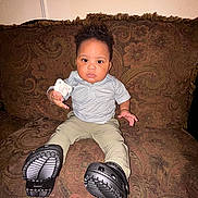 Rory is registered to the contest to win money with this photo: toddler, child, pacifier, couch, indoor, footwear, patterned_fabric, sitting, serious_expression, short_sleeves, pants, black_shoes, hand, person, home, young_child, curly_hair, brown_couch, furniture, portrait