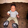 toddler, child, pacifier, couch, indoor, footwear, patterned_fabric, sitting, serious_expression, short_sleeves, pants, black_shoes, hand, person, home, young_child, curly_hair, brown_couch, furniture, portrait