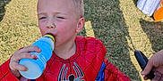 Samuel is registered to the contest to win money with this photo: child, boy, spiderman_costume, drinking, bottle, outdoor, grass, sunlight, shadow, hand, costume, playful, daylight, casual, fun, person, footwear, shorts, holding, summer