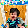 toddler, child, smiling, happy, striped_shirt, high_chair, indoor, door, wooden_door, black_hair, big_eyes, face, person, baby, clothing, seat, colorful, banner, stars, playful