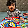toddler, child, pajamas, bed, puzzle, toy, thomas_and_friends, blue, red, hand, face, smiling, indoors, headboard, curly_hair, playtime, person, young_child, furniture, happy