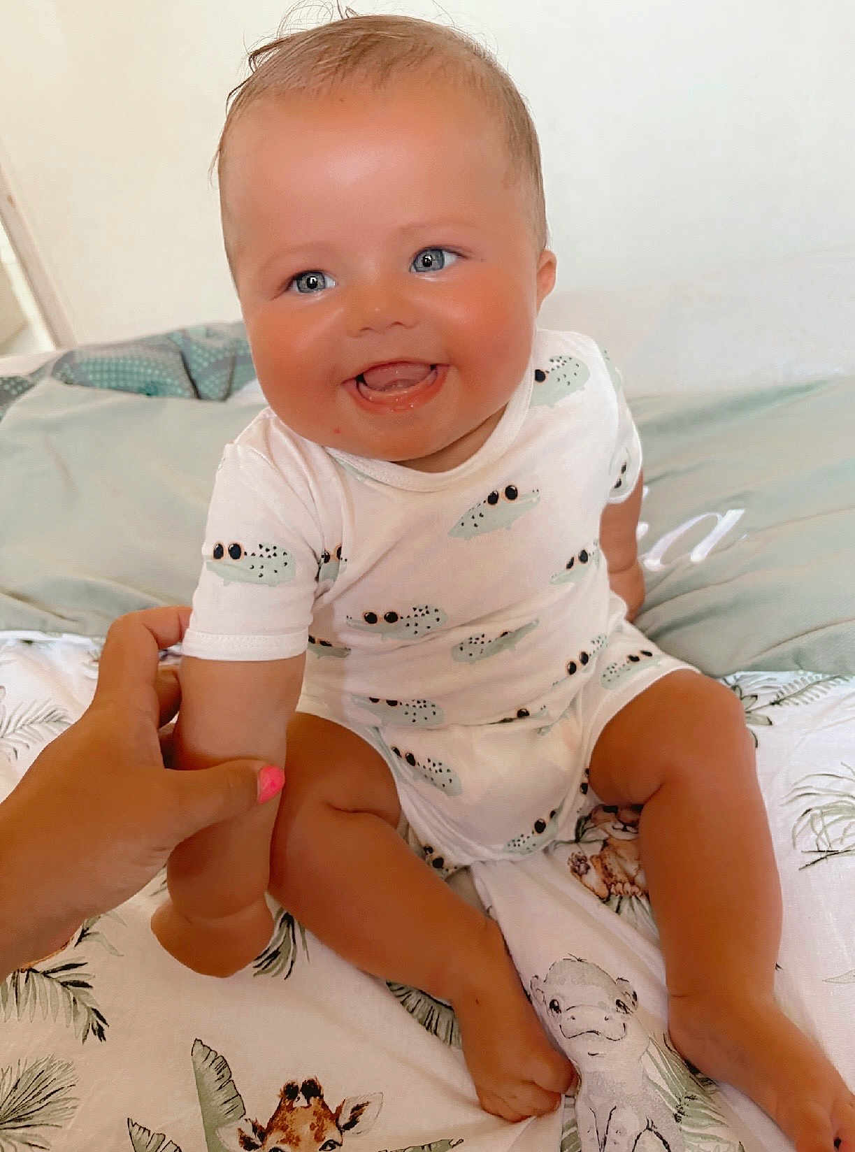 Sacha participe au concours pour gagner de l'argent avec cette photo : baby, child, smiling, blue_eyes, white_onesie, crocodile_pattern, bed, animal_prints, hand, holding, cute, happy, infant, person, skin, sitting, indoors, comfort, care, portrait