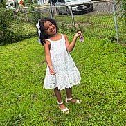 Anaya is registered to the contest to win money with this photo: child, girl, smiling, outdoor, portrait, white_dress, lace_dress, long_hair, hair_bow, sandals, grass, lawn, garden, chain_link_fence, residential_area, houses, parked_cars, street, sunlight, happy