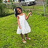 child, girl, smiling, outdoor, portrait, white_dress, lace_dress, long_hair, hair_bow, sandals, grass, lawn, garden, chain_link_fence, residential_area, houses, parked_cars, street, sunlight, happy