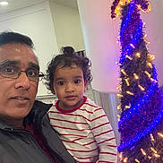 Juliet Mariam joined the competition — help win amazing prizes! adult, child, toddler, striped_shirt, glasses, indoor, christmas_tree, lights, holiday_decor, curly_hair, person, family, portrait, casual_clothing, smile, holding, festive, celebration, happy, together