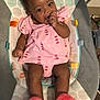 baby, child, infant, pink_dress, watermelon_print, slippers, baby_rocker, indoor, carpet, sitting, thumb_sucking, cute, smiling, happy, person, furniture, footwear, patterned, colorful, comfortable