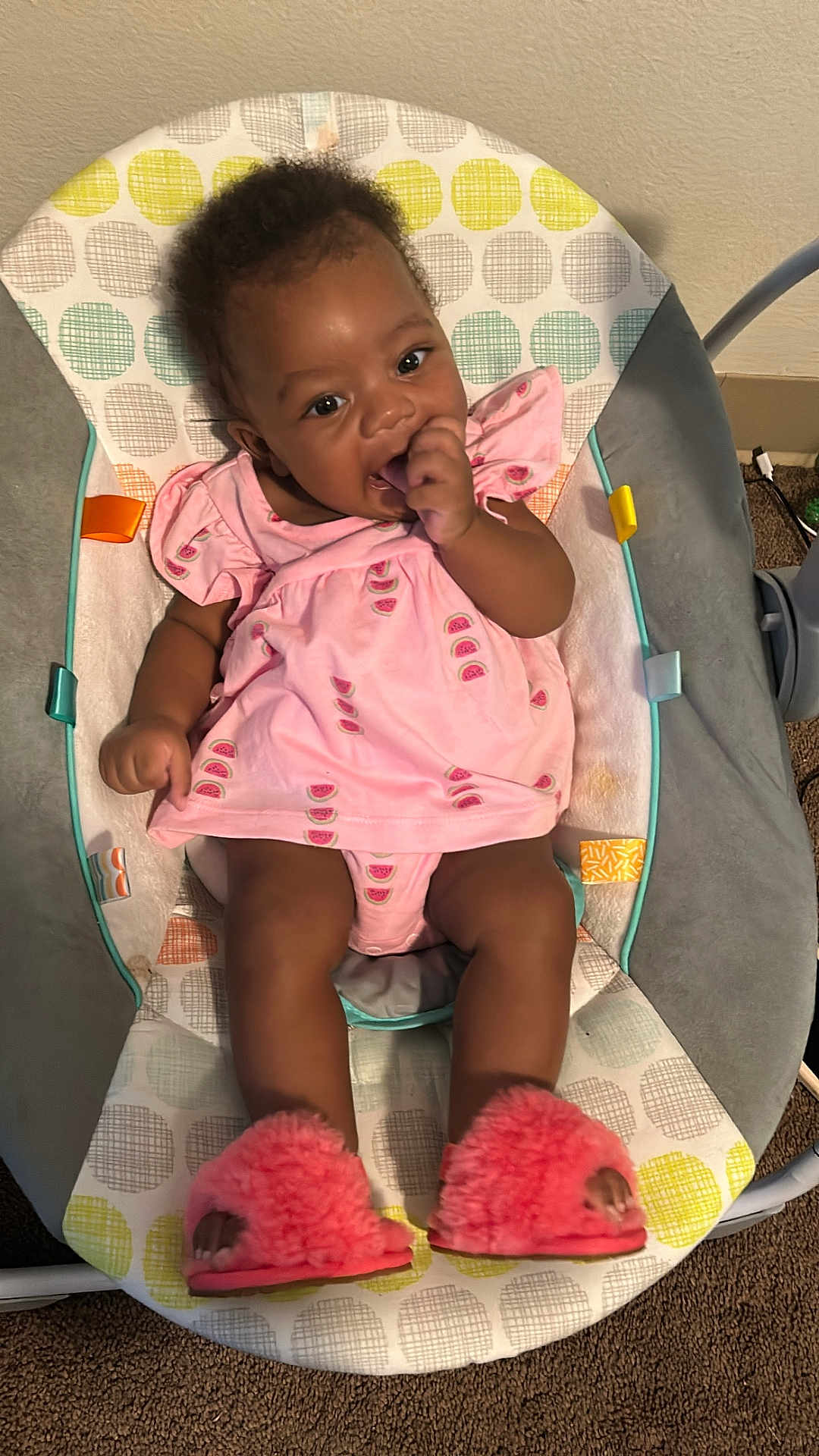 Anari is registered to the contest to win money with this photo: baby, child, infant, pink_dress, watermelon_print, slippers, baby_rocker, indoor, carpet, sitting, thumb_sucking, cute, smiling, happy, person, furniture, footwear, patterned, colorful, comfortable