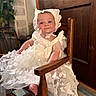 adorable, baby, bonnet, butterfly_pattern, carpet, child, face, formal_attire, furniture, indoor, infant, lace, photography, portrait, ribbon, rocking_chair, sitting, smiling, white_dress, wooden_door
