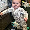 baby, baby_wipes, blanket, carpet, child, clothing, couch, face, floor, furniture, hand, head, indoor, messy_room, onesie, person, sheep_pattern, smiling, standing, toy