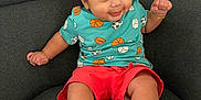 Reyaansh is registered to the contest to win money with this photo: baby, infant, child, smiling, sitting, armchair, couch, teal_shirt, coral_shorts, sports_pattern, barefoot, toes, bracelet, dark_hair, big_eyes, portrait, indoors, cute, happy, person