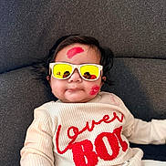 Reyaansh is registered to the contest to win money with this photo: baby, infant, sunglasses, lipstick, kiss_mark, white_outfit, red_text, couch, gray_background, lying_down, portrait, chubby_cheeks, playful, closeup, reflection, indoors, smile, cute, family, clothing