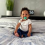 baby, pacifier, bed, bedroom, plant, clock, smiling, white_shirt, blue_pants, curly_hair, indoor, child, person, furniture, modern, home, cute, sitting, portrait, happy